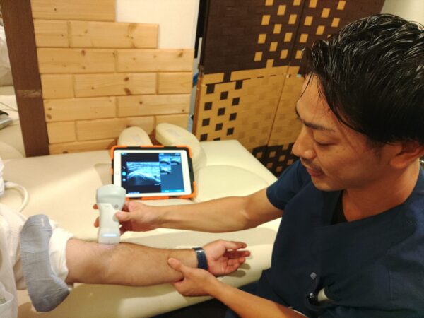 施術者が、患者の肘から前腕にかけてエコー検査（超音波検査）を行っており、iPadの画面に画像が映し出されている。