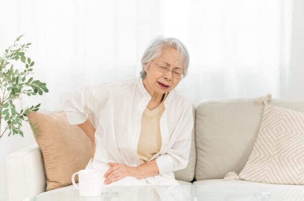 ソファに座った高齢の女性が、腰と首を抑えて痛みに顔をゆがめている。手元にはマグカップがある。