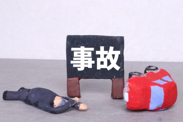 「事故」と書かれた黒いミニ看板、倒れている人形、横転した赤いミニカーが置かれている。