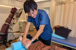 横須賀市の鍼灸整骨院ひまわりで、施術者がアキレス腱炎の患者の足首とふくらはぎに手技施術を行っている様子。奥にはトムソンテーブルが見える。
