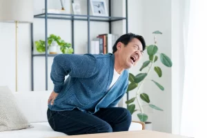 自宅のソファで突然のぎっくり腰による激しい痛みに苦しんでいる男性の画像。横須賀市の鍼灸整骨院ひまわりはぎっくり腰に緊急対応。