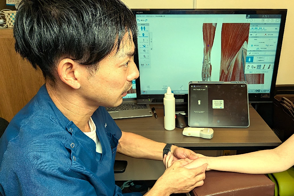 横須賀の鍼灸整骨院ひまわりで、施術者がTFCC損傷が疑われる患者の手首の小指側を丁寧に触診し、モニターで手首の構造を説明している様子